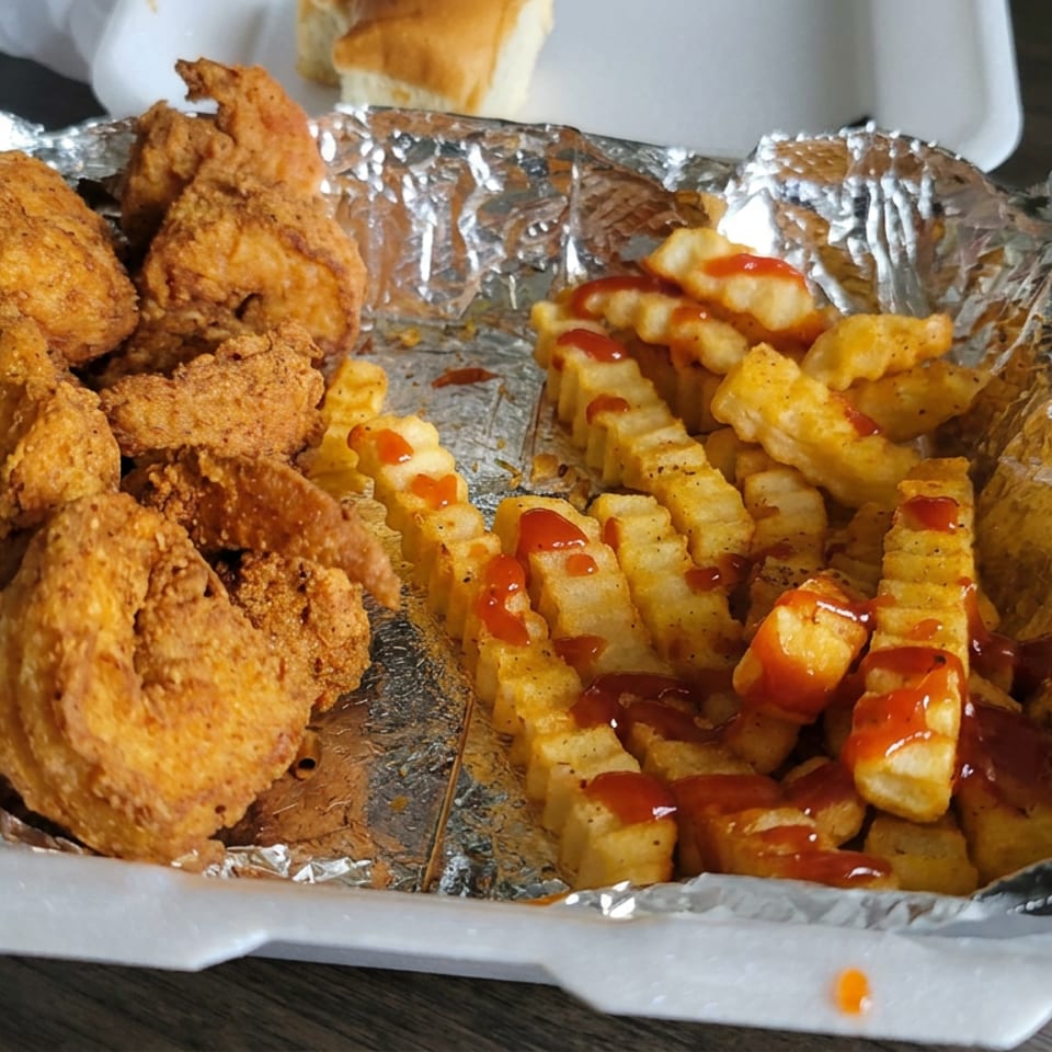Takeout container holding crispy fried chicken pieces and seasoned crinkle-cut fries drizzled with ketchup.