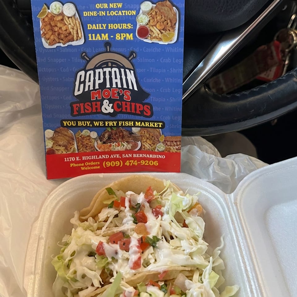 Open takeout container holding fish tacos topped with shredded cabbage, tomatoes, and creamy sauce, beside a Captain Moe’s Fish & Chips flyer.