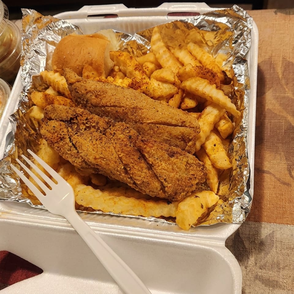 Takeout container filled with fried fish, crinkle-cut fries, hush puppies, and side sauces on a patterned tablecloth.