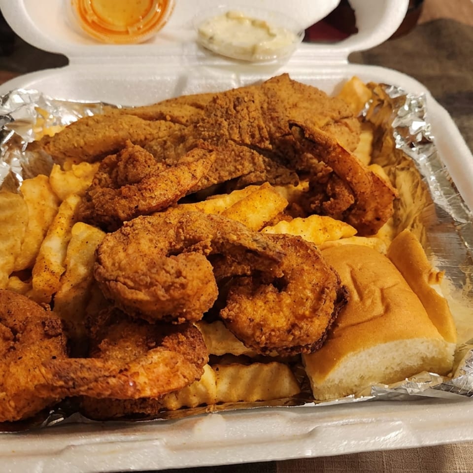 Foam container filled with fried seafood, macaroni and cheese, fries, and a dinner roll, with dipping sauces beside it.